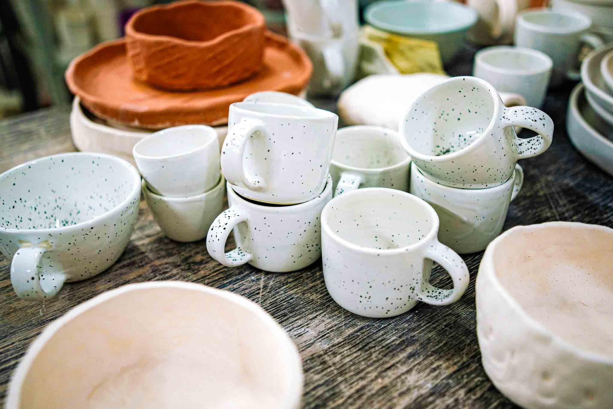 Collection of handcrafted pottery cups and bowls in white with black speckles and terracotta plates stacked on a table as part of pottery making experience in Yelahanka.